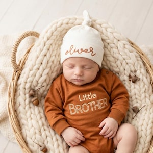 Può includere: Un body a maniche lunghe color ruggine con la scritta Little BROTHER ricamata in bianco e un berretto annodato color crema abbinato con il nome oliver ricamato in ruggine, indossato da un neonato che dorme in un cesto intrecciato.