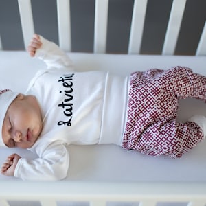 May include: A baby wearing a white long-sleeved onesie with the word "Latvieti" printed on it, a matching burgundy and white patterned hat, and burgundy and white patterned pants. The baby is sleeping in a white crib.