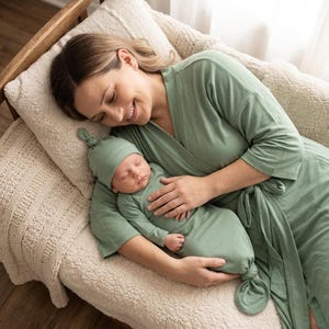 Puede incluir: Una mujer y un bebé están acurrucados juntos sobre una almohada color crema. La mujer lleva una bata verde salvia, y el bebé está con un vestido y gorro anudados a juego. La escena evoca una sensación de comodidad y tranquilidad.
