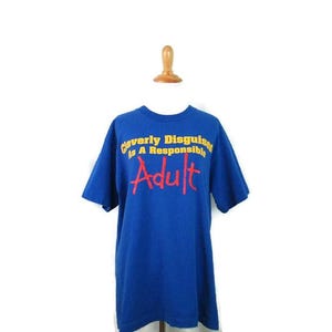 May include: A blue t-shirt with the text "Cleverly Disguised as a Responsible Adult" in yellow and red lettering. The shirt is displayed on a wooden mannequin head and torso against a white background.