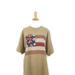 Puede incluir: Una camiseta de manga corta de color marr&oacute;n claro con un dise&ntilde;o de bandera americana de patchwork con estrellas rojas, blancas y azules.