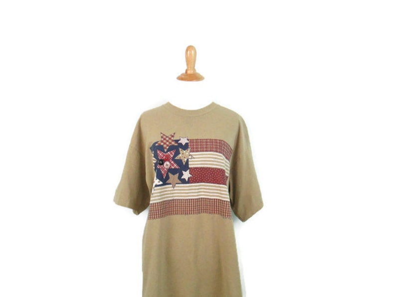 Puede incluir: Una camiseta de manga corta de color marr&oacute;n claro con un dise&ntilde;o de bandera americana de patchwork con estrellas rojas, blancas y azules.