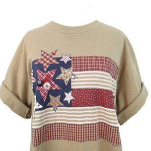 Puede incluir: Una camiseta de color marr&oacute;n claro con un dise&ntilde;o de bandera americana de patchwork con estrellas rojas, blancas y azules y botones. La bandera est&aacute; hecha de diferentes telas, incluyendo cuadros y rayas.