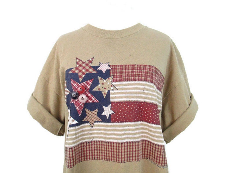 Puede incluir: Una camiseta de color marr&oacute;n claro con un dise&ntilde;o de bandera americana de patchwork con estrellas rojas, blancas y azules y botones. La bandera est&aacute; hecha de diferentes telas, incluyendo cuadros y rayas.