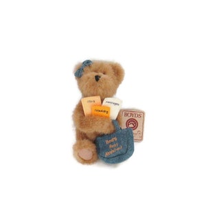 May include: A brown teddy bear wearing a blue denim bag with the text "Bear-y Best Assistant" on it. The bear is holding several small orange cards with text on them. The bear is also holding a small brown tag with the text "BOYDS" and a paw print.