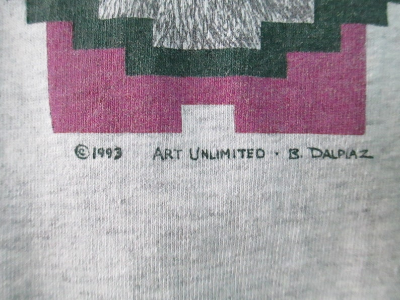 Op de afbeelding: Een grijs T-shirt met een geometrisch ontwerp in roze, groen en grijs. De tekst "&copy; 1993 ART UNLIMITED - B. DALPIAZ" staat onderaan het shirt gedrukt.