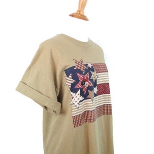 Puede incluir: Una camiseta de manga corta de color marr&oacute;n claro con un dise&ntilde;o de patchwork con estrellas y rayas rojas, blancas y azules. El dise&ntilde;o recuerda a una bandera estadounidense.