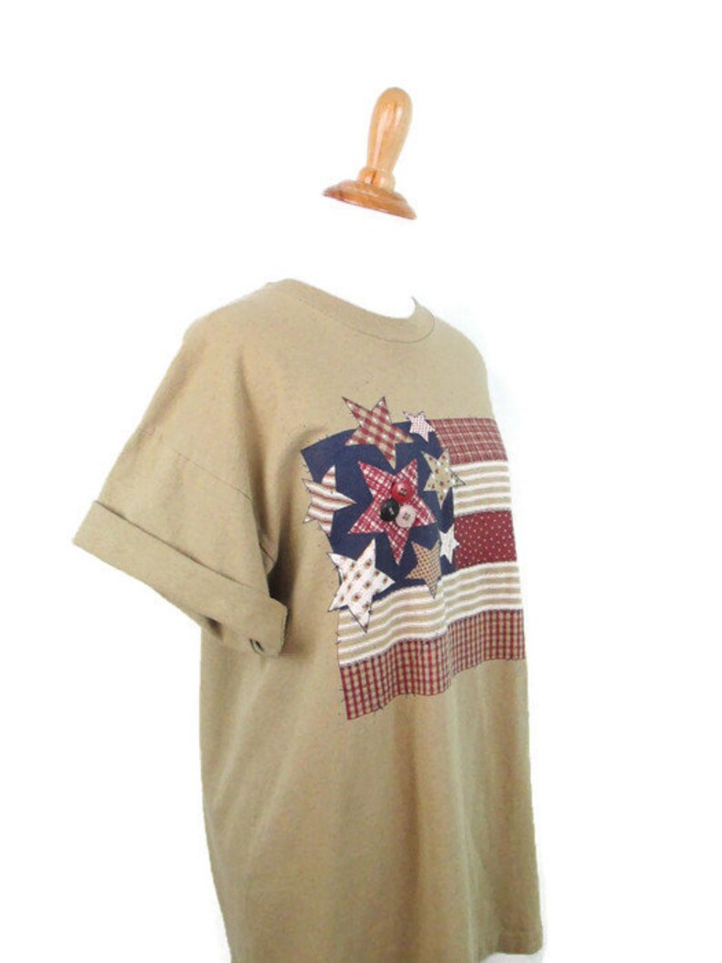 Puede incluir: Una camiseta de manga corta de color marr&oacute;n claro con un dise&ntilde;o de patchwork con estrellas y rayas rojas, blancas y azules. El dise&ntilde;o recuerda a una bandera estadounidense.