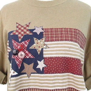 Puede incluir: Camiseta de color marr&oacute;n claro con un dise&ntilde;o de bandera americana de patchwork con estrellas rojas, blancas y azules y un patr&oacute;n de cuadros.