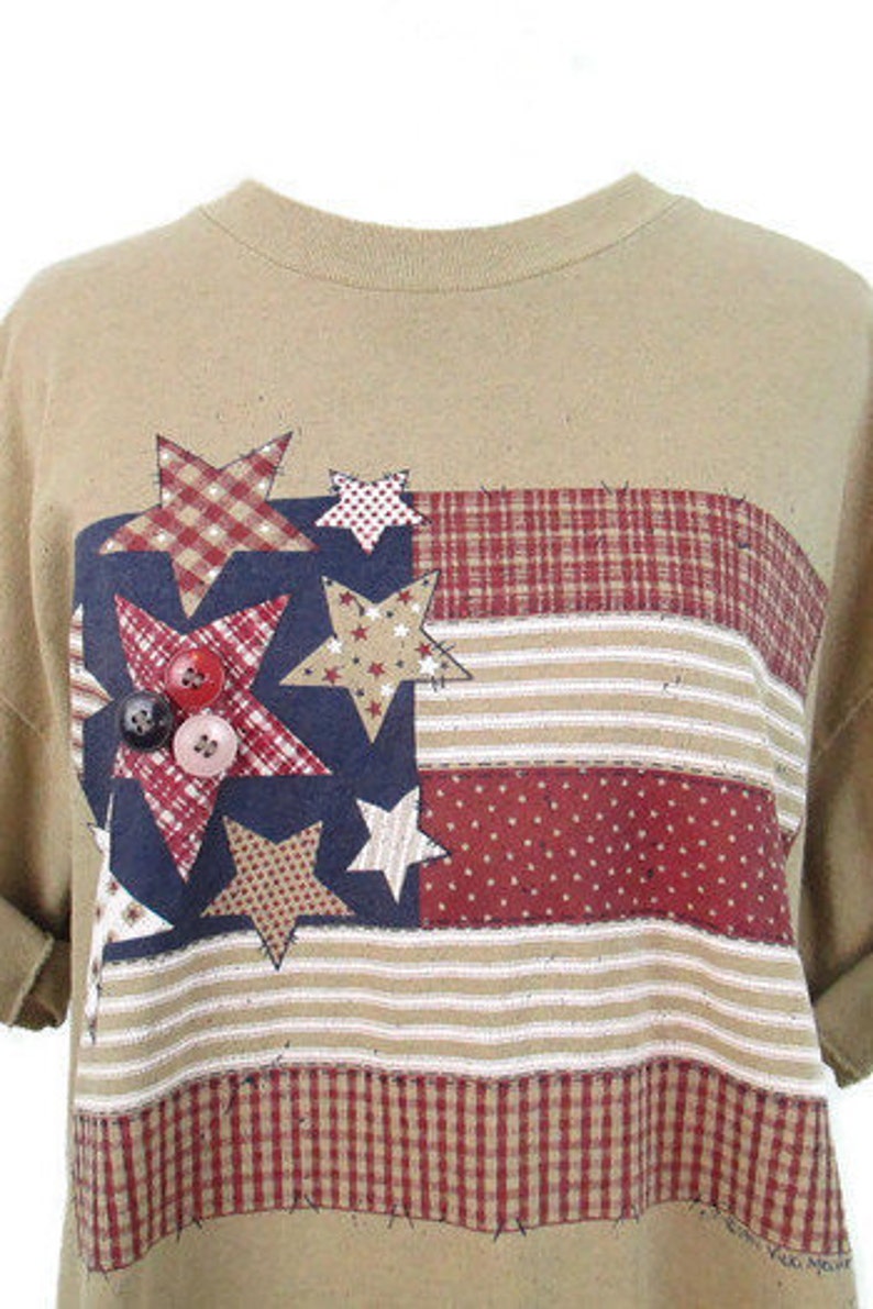 Puede incluir: Camiseta de color marr&oacute;n claro con un dise&ntilde;o de bandera americana de patchwork con estrellas rojas, blancas y azules y un patr&oacute;n de cuadros.