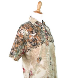 May include: A beige short-sleeved button-up shirt with a lion print. The shirt has a faded look and features a brown, black, and red color scheme.