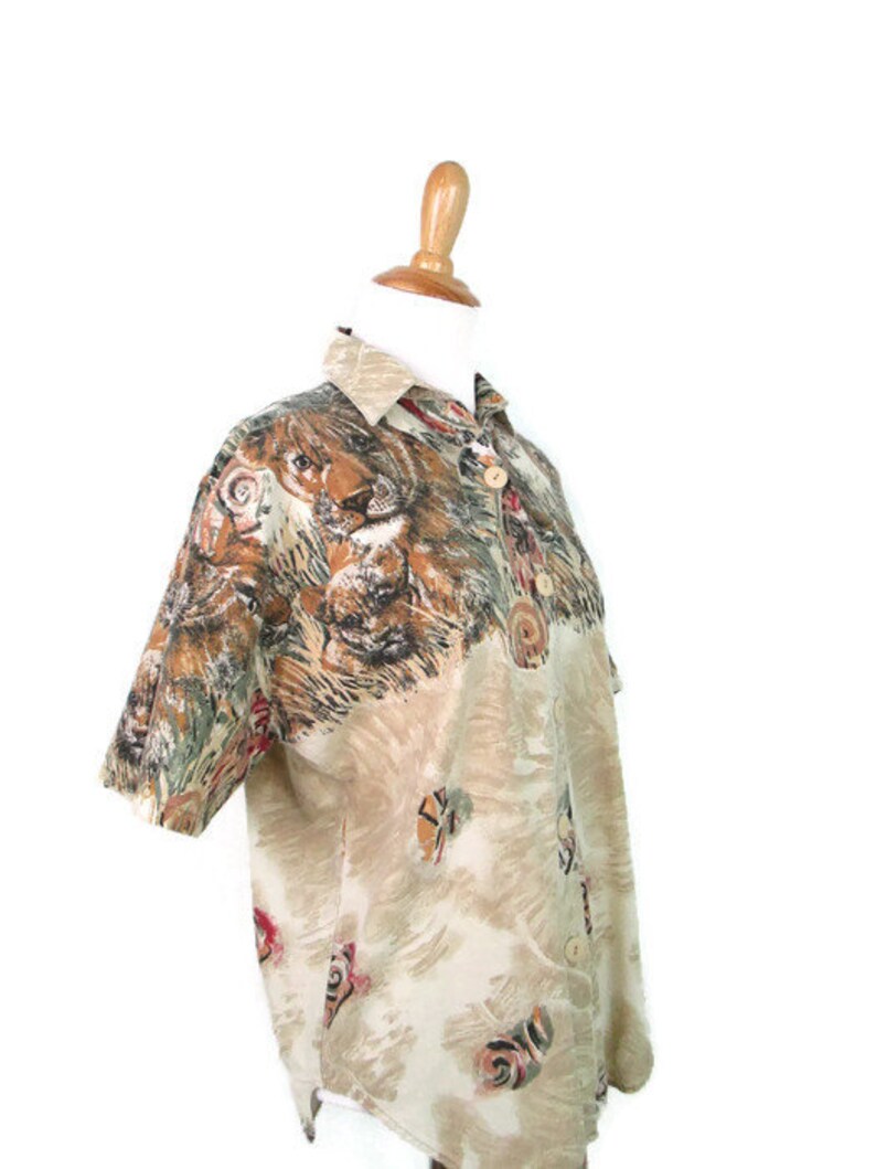 May include: A beige short-sleeved button-up shirt with a lion print. The shirt has a faded look and features a brown, black, and red color scheme.