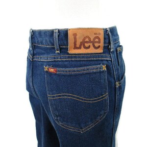 Pode incluir: Um par de cal&ccedil;as de ganga azul escuro com um remendo de couro castanho na parte de tr&aacute;s da cintura que diz "Lee" em letras castanhas. As cal&ccedil;as t&ecirc;m um bolso traseiro com um pequeno logotipo Lee vermelho.
