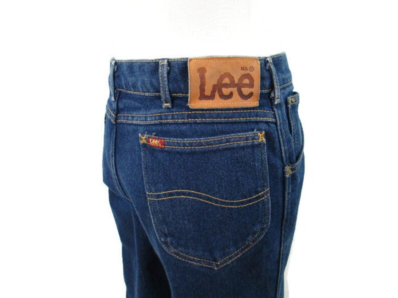 Pode incluir: Um par de cal&ccedil;as de ganga azul escuro com um remendo de couro castanho na parte de tr&aacute;s da cintura que diz "Lee" em letras castanhas. As cal&ccedil;as t&ecirc;m um bolso traseiro com um pequeno logotipo Lee vermelho.
