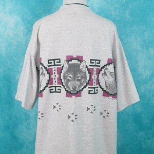 Op de afbeelding: Grijs T-shirt met korte mouwen met een wolfprint. De wolf is omringd door een geometrisch patroon in tinten groen, roze en wit. De onderkant van het shirt heeft een spoor van wolfspoten.