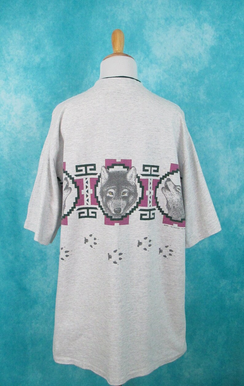 Op de afbeelding: Grijs T-shirt met korte mouwen met een wolfprint. De wolf is omringd door een geometrisch patroon in tinten groen, roze en wit. De onderkant van het shirt heeft een spoor van wolfspoten.