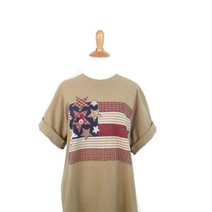 Puede incluir: Una camiseta de manga corta de color marr&oacute;n claro con un dise&ntilde;o de bandera americana de patchwork con estrellas y rayas rojas, blancas y azules.