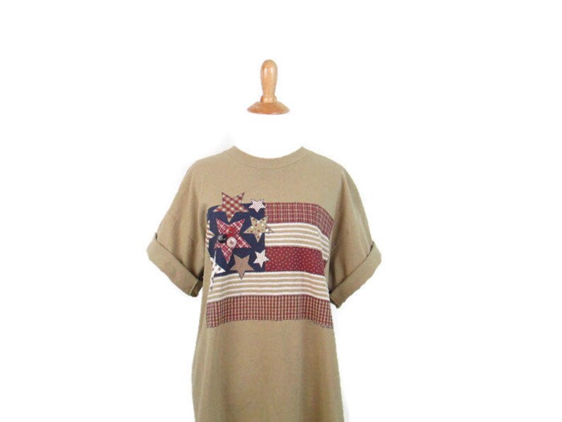 Puede incluir: Una camiseta de manga corta de color marr&oacute;n claro con un dise&ntilde;o de bandera americana de patchwork con estrellas y rayas rojas, blancas y azules.