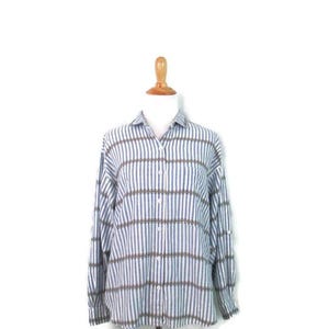 Puede incluir: Camisa a rayas verticales blancas, azules y marrones, exhibida en un maniquí de madera. La camisa de manga larga tiene un cuello clásico y puños abotonados. El fondo es blanco liso. La tela es ligera.