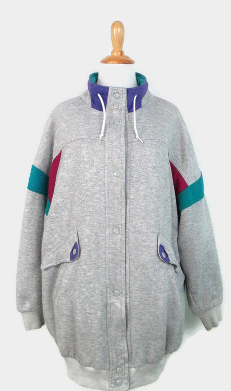 May include: A gray sweatshirt jacket with a snap closure and a high collar. The jacket has purple and teal colored stripes on the sleeves.