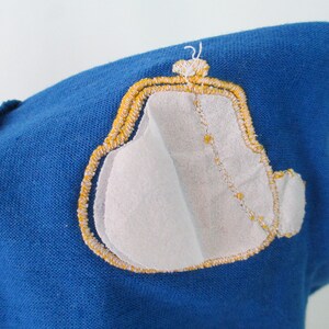 May include: A blue fabric patch with a yellow embroidered design of a purse. The purse is outlined in yellow and has a white fabric interior.