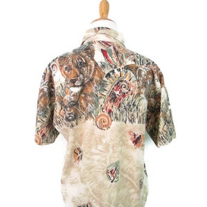 May include: A beige button-up shirt with a short sleeve. The shirt has a colorful print of a lion, feathers, and other abstract designs.