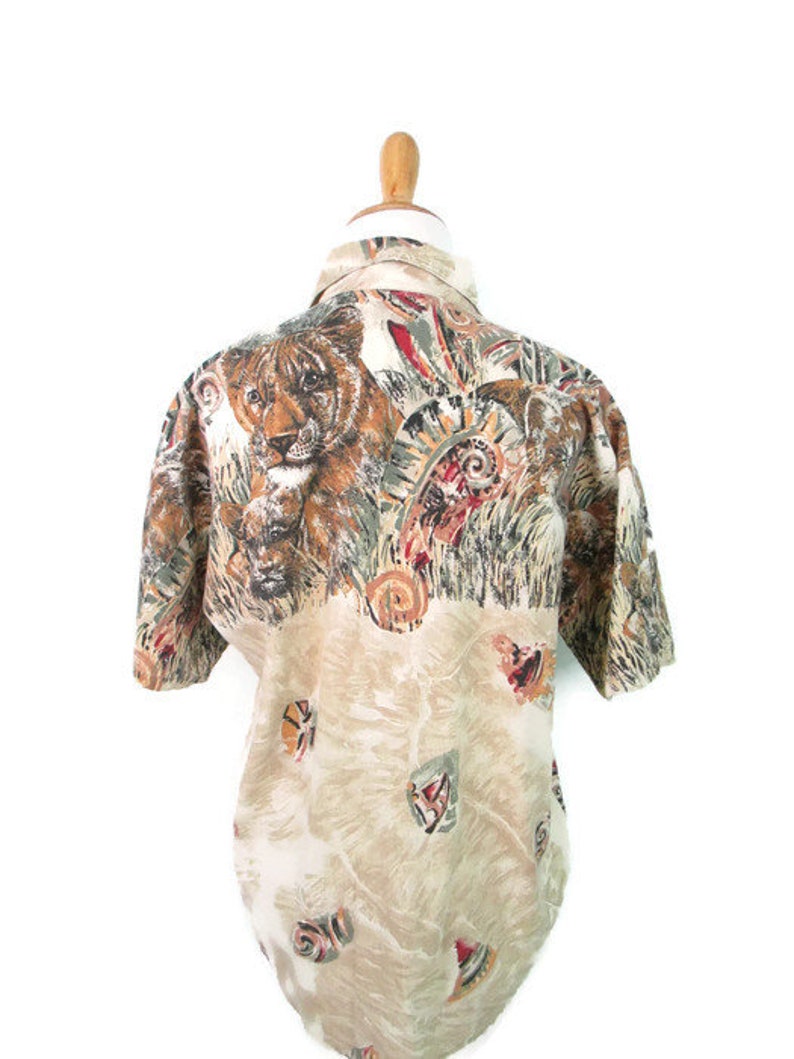 May include: A beige button-up shirt with a short sleeve. The shirt has a colorful print of a lion, feathers, and other abstract designs.