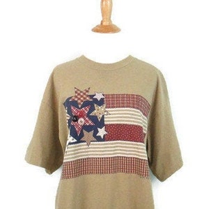 Puede incluir: Una camiseta de manga corta de color marr&oacute;n claro con un dise&ntilde;o de bandera americana de patchwork con estrellas y rayas rojas, blancas y azules.