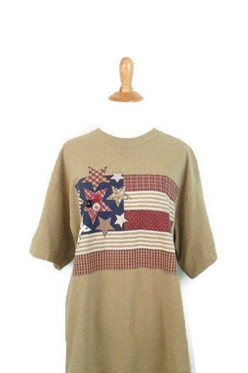 Puede incluir: Una camiseta de manga corta de color marr&oacute;n claro con un dise&ntilde;o de bandera americana de patchwork con estrellas y rayas rojas, blancas y azules.