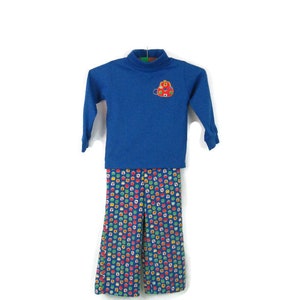 May include: A blue long-sleeved turtleneck shirt with a red and yellow embroidered backpack design. The shirt is paired with blue pants with a colorful geometric pattern.