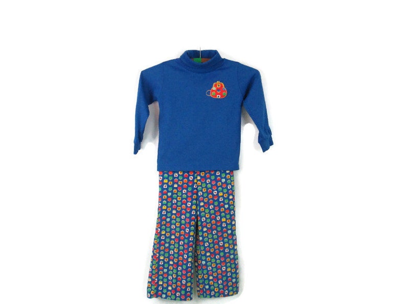May include: A blue long-sleeved turtleneck shirt with a red and yellow embroidered backpack design. The shirt is paired with blue pants with a colorful geometric pattern.