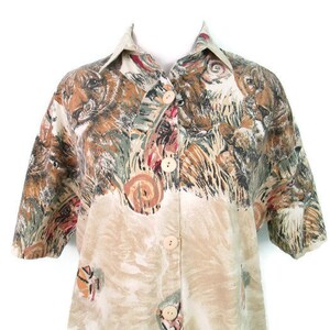 May include: A beige button-up shirt with a short sleeve and a colorful animal print. The print features a variety of animals, including tigers, leopards, and other wild cats.