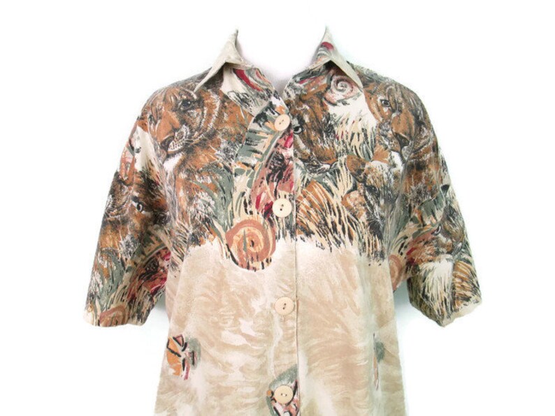 May include: A beige button-up shirt with a short sleeve and a colorful animal print. The print features a variety of animals, including tigers, leopards, and other wild cats.