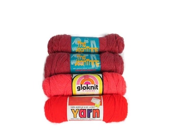 May include: Four skeins of yarn in shades of red, stacked vertically against a white background. The top two skeins are a deep red with blue labels that read "The Weaving Time." The third skein is a bright coral with a "gloknit" label. The bottom skein is a vibrant red with a "Yarn" label.