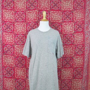 May include: A gray t-shirt with a single pocket on the left side. The shirt is hanging on a mannequin against a red and white patterned background.