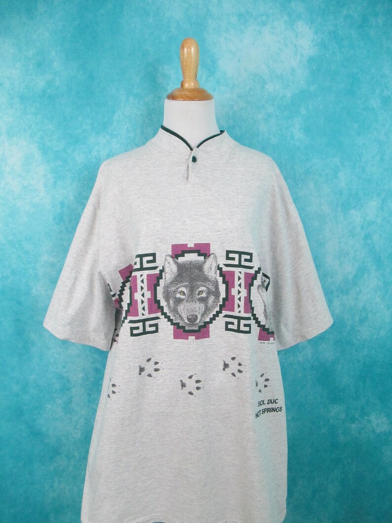 Op de afbeelding: Grijs T-shirt met korte mouwen met een wolf-print en voetafdrukken. Het shirt heeft een mandarijnskraag en een knoopsluiting. De tekst "Sol Duc Hot Springs" staat gedrukt op de rechteronderkant van het shirt.