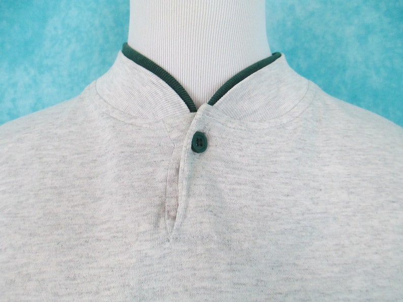 Op de afbeelding: Een grijs longsleeve shirt met een groene kraag en een enkele knoopsluiting aan de hals.