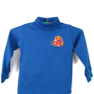 May include: A blue long-sleeved turtleneck shirt with an embroidered red purse with colorful details on the front.
