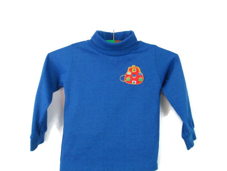 May include: A blue long-sleeved turtleneck shirt with an embroidered red purse with colorful details on the front.
