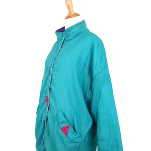 May include: A teal blue windbreaker jacket with pink triangle accents on the pockets. The jacket has a zipper closure and a stand-up collar.