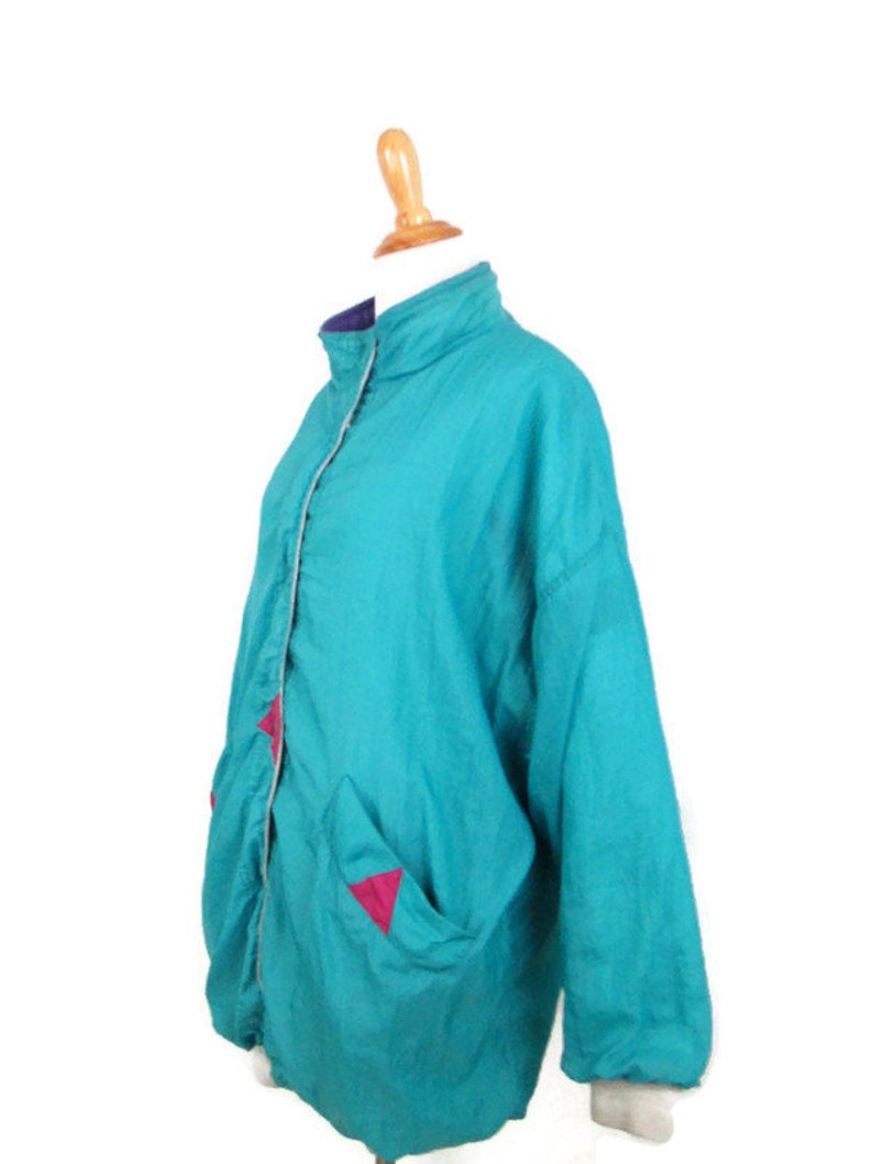 May include: A teal blue windbreaker jacket with pink triangle accents on the pockets. The jacket has a zipper closure and a stand-up collar.