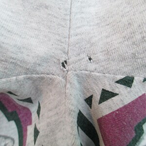 Op de afbeelding: Close-up van een grijs T-shirt met twee kleine gaatjes in de buurt van de naad. Het shirt heeft een kleurrijke grafische print met geometrische vormen en een paars-groen ontwerp.