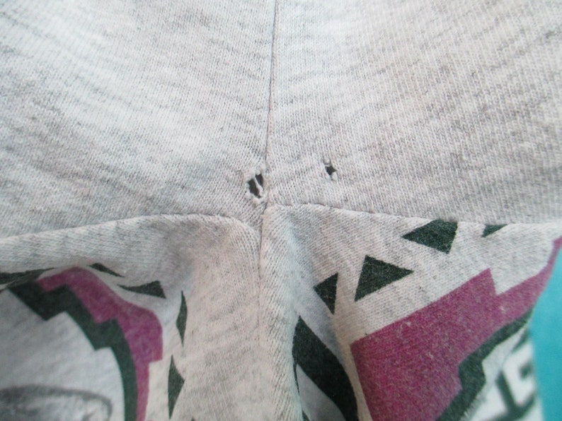 Op de afbeelding: Close-up van een grijs T-shirt met twee kleine gaatjes in de buurt van de naad. Het shirt heeft een kleurrijke grafische print met geometrische vormen en een paars-groen ontwerp.