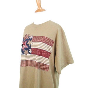 Puede incluir: Una camiseta de manga corta de color marr&oacute;n claro con un dise&ntilde;o de bandera americana de patchwork con estrellas y rayas rojas, blancas y azules.