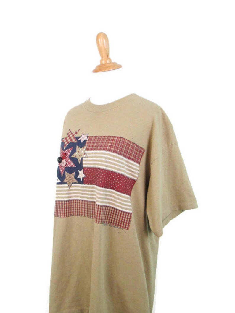 Puede incluir: Una camiseta de manga corta de color marr&oacute;n claro con un dise&ntilde;o de bandera americana de patchwork con estrellas y rayas rojas, blancas y azules.