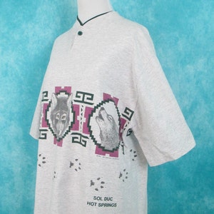 Op de afbeelding: Een grijs T-shirt met korte mouwen met een grafische print van drie wolfskopjes in een Native American stijl. Het shirt heeft een knoopsluiting en de tekst "Sol Duc Hot Springs" staat onderaan gedrukt.
