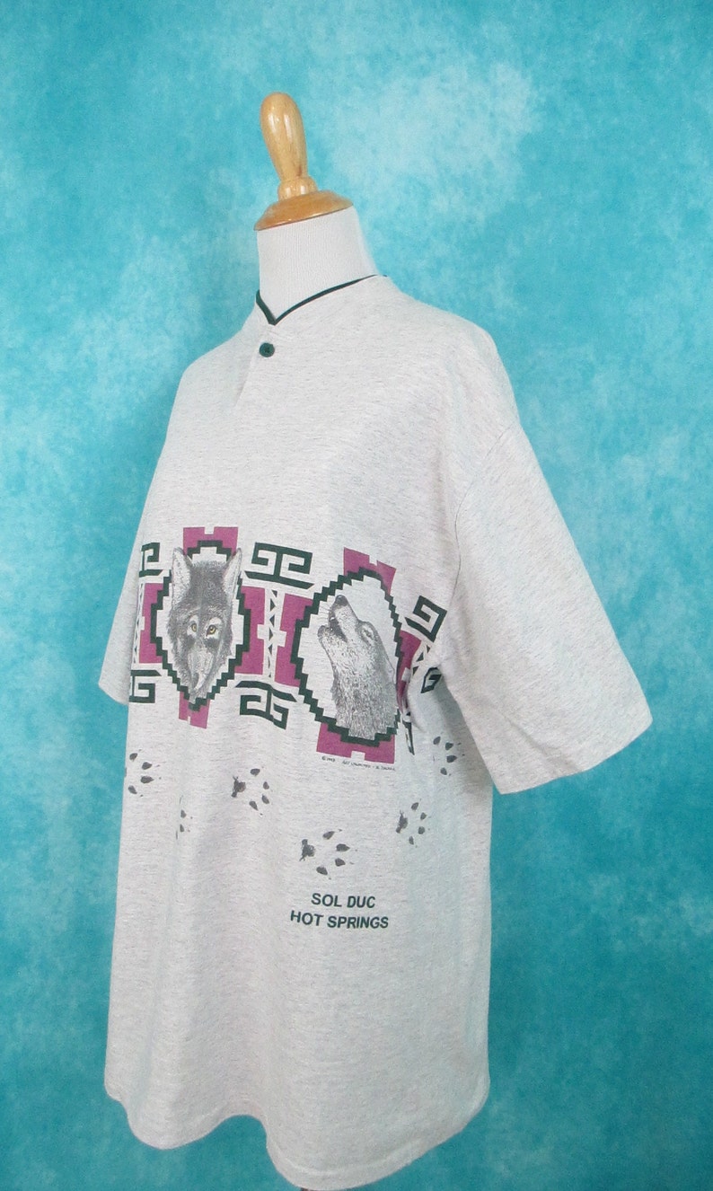 Op de afbeelding: Een grijs T-shirt met korte mouwen met een grafische print van drie wolfskopjes in een Native American stijl. Het shirt heeft een knoopsluiting en de tekst "Sol Duc Hot Springs" staat onderaan gedrukt.