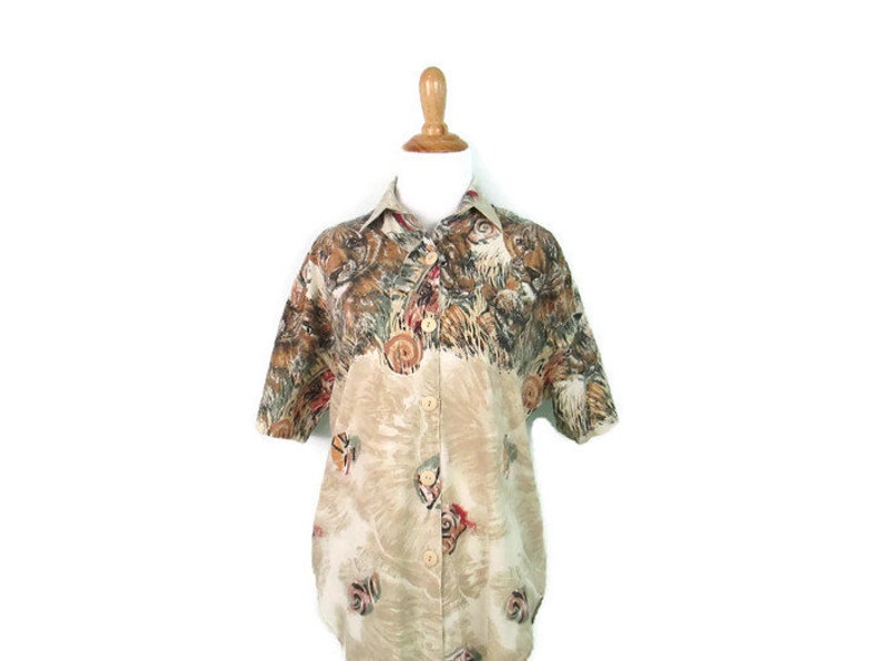May include: A beige short-sleeved button-down shirt with a colorful print of animals. The print features a variety of animals, including tigers, leopards, and other wildlife.