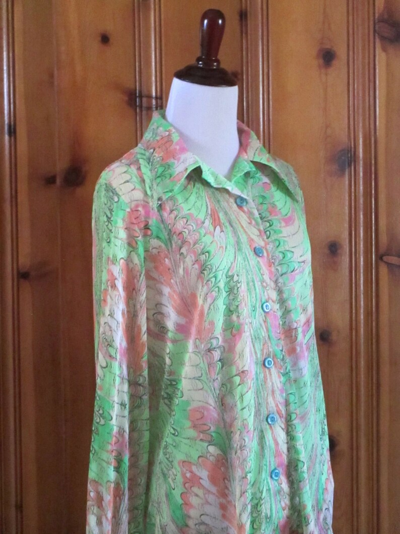 May include: A long-sleeved, green and pink patterned shirt with a collared neckline and blue buttons. The shirt is made of a lightweight fabric and has a flowing silhouette.