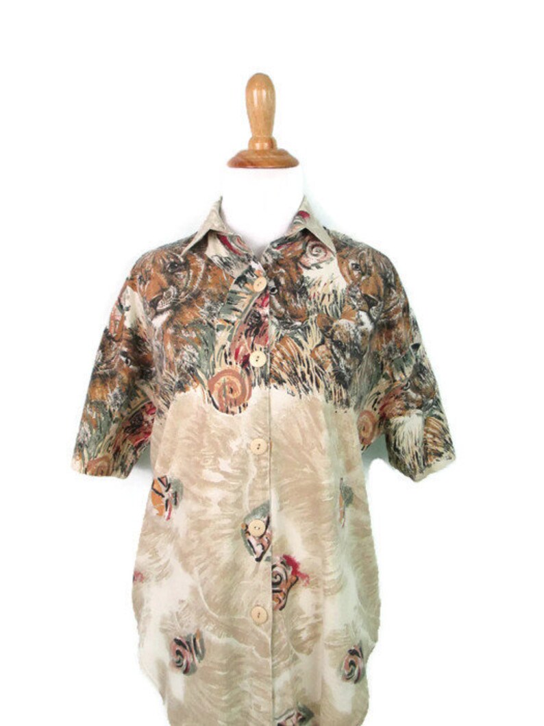 May include: A beige button-up shirt with a short sleeve and a colorful animal print. The print features a variety of animals, including lions, tigers, and leopards.